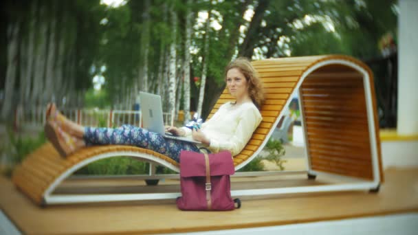Mooie vrouw eet een ijsje en werkt op een laptop op een houten bankje in het park — Stockvideo