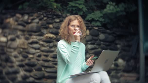 Woman with a laptop and documents on the background of a stony wall. Drinks and drinks wine from a glass — Stock Video