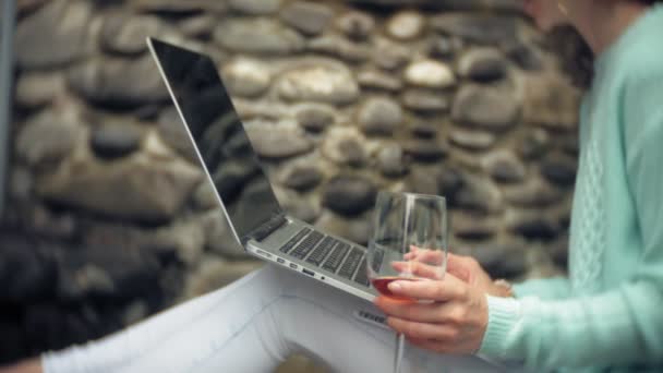 Kvinna med en laptop och dokument på bakgrunden av en stenig vägg. Drycker och drinkar vin ur ett glas — Stockvideo