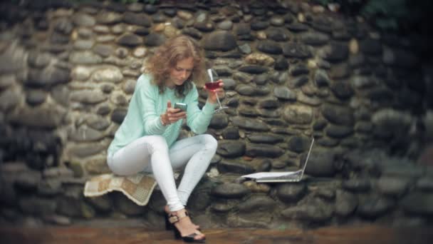 Frau mit Laptop und Dokumenten vor einer steinernen Mauer. Getränke und Wein aus einem Glas — Stockvideo