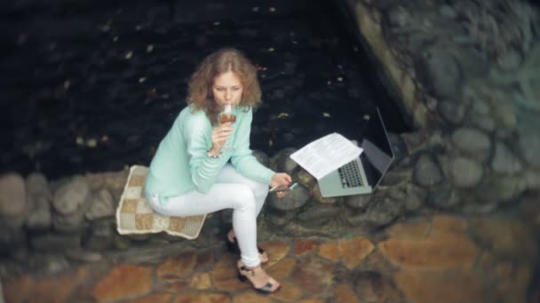 Donna con un computer portatile e documenti sullo sfondo di un muro di pietra. Bevande e vino di bevande da un bicchiere — Video Stock