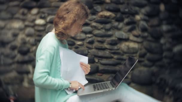 Mulher com um laptop e documentos no fundo de uma parede de pedra . — Vídeo de Stock