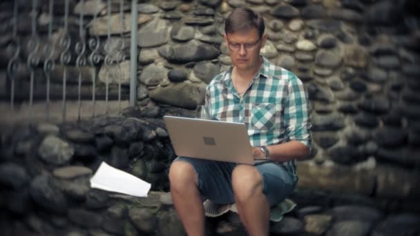 Ein Mann mit Laptop und Dokumenten auf dem Hintergrund einer Steinmauer arbeitet und trinkt Wein aus einem Glas — Stockvideo