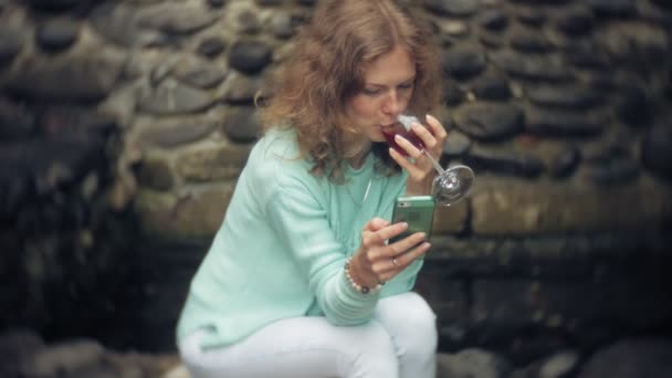 Uma mulher está usando um smartphone contra uma parede traseira. Bebidas e bebidas vinho de um copo — Vídeo de Stock