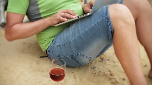 Busy mature man working on a laptop computer with business papers picking up a phone to work a call sitting on the beach by the sea on a sunny day and drinking wine from a glass — Stock Video