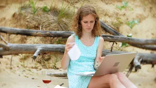 Hermosa mujer joven ocupada trabajando en una computadora portátil con papeles de negocios recogiendo un teléfono para trabajar una llamada sentada en la playa junto al mar en un día soleado y bebiendo vino de una copa — Vídeos de Stock