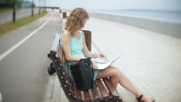 Жінка сидить на лавці на пляжі, використовуючи ноутбук — стокове відео