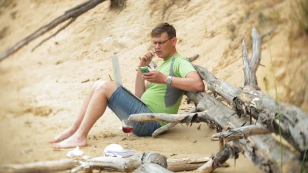 Upptagen mogen man arbetar på en bärbar dator med affärshandlingar plocka upp en telefon att arbeta ett samtal som sitter på stranden vid havet på en solig dag och dricka vin ur ett glas — Stockvideo