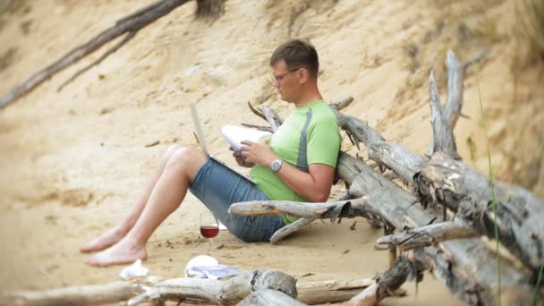 Beschäftigter älterer Mann, der an einem Laptop arbeitet, auf dem Geschäftspapiere liegen und zum Telefon greift, um an einem sonnigen Tag am Strand am Meer zu telefonieren und Wein aus einem Glas zu trinken — Stockvideo