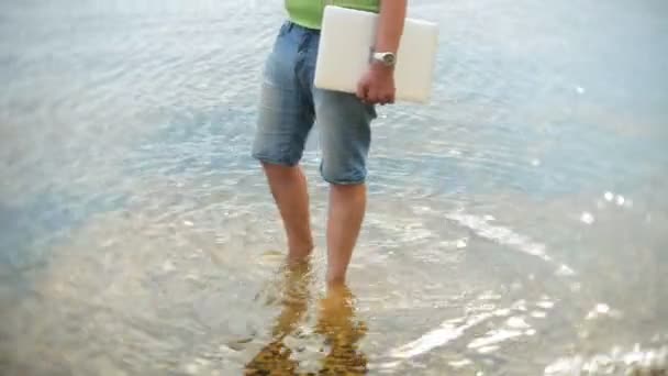 Ένας άνθρωπος στέκεται στο νερό με έναν φορητό υπολογιστή στην παραλία το καλοκαίρι — Αρχείο Βίντεο