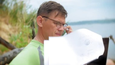 bir telefon bir güneşli gün ve içme şarap cam sahilde denizin kenarında oturan bir çağrı için malzeme çekme iş kağıtları ile bir dizüstü bilgisayarda çalışma meşgul olgun adam