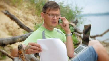 bir telefon bir güneşli gün ve içme şarap cam sahilde denizin kenarında oturan bir çağrı için malzeme çekme iş kağıtları ile bir dizüstü bilgisayarda çalışma meşgul olgun adam
