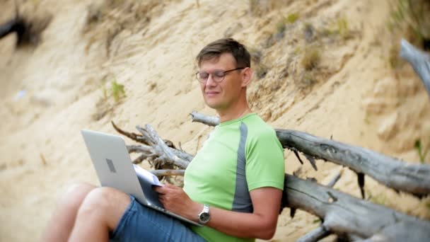 Занятой взрослый человек, работающий за ноутбуком с деловыми бумагами, берущий трубку телефона для работы, сидящий на пляже у моря в солнечный день и пьющий вино из бокала — стоковое видео