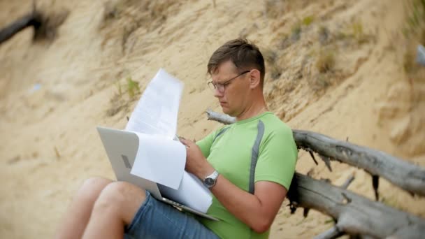 Beschäftigter älterer Mann, der an einem Laptop arbeitet, auf dem Geschäftspapiere liegen und zum Telefon greift, um an einem sonnigen Tag am Strand am Meer zu telefonieren und Wein aus einem Glas zu trinken — Stockvideo