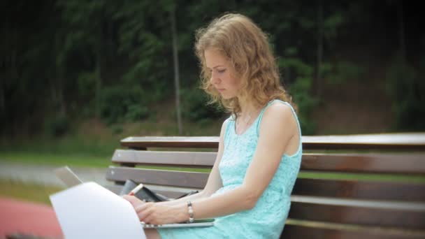 Una mujer sentada en un banco en la playa disfruta de un portátil en un documento de lectura — Vídeos de Stock