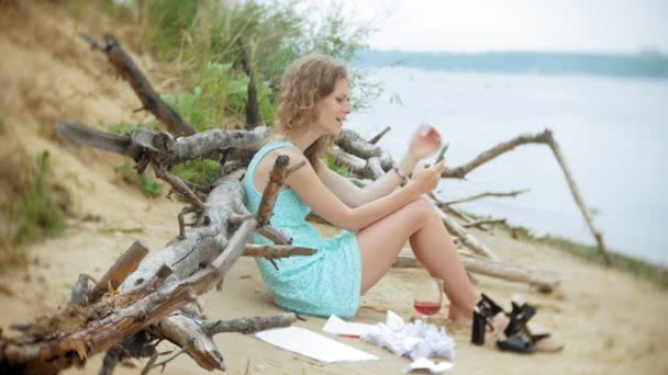 Busy beautiful young woman working on a laptop computer with business papers picking up a phone to work a call sitting on the beach by the sea on a sunny day and drinking wine from a glass — Stock Video