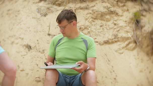 Busy mature man working on a laptop computer with business papers picking up a phone to work a call sitting on the beach by the sea on a sunny day and drinking wine from a glass — Stock Video