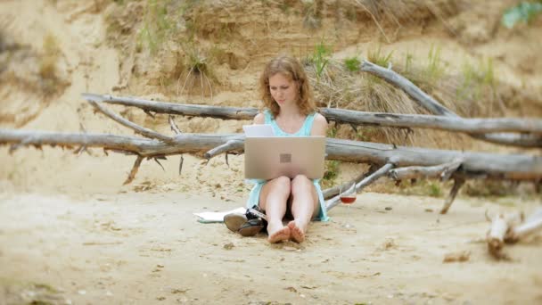 Απασχολημένος όμορφη νεαρή γυναίκα που εργάζεται σε ένα φορητό υπολογιστή με επαγγελματικά έγγραφα μαζεύοντας ένα τηλέφωνο για να λειτουργήσει μια κλήση που κάθονται στην παραλία δίπλα στη θάλασσα, σε μια ηλιόλουστη ημέρα και πίνοντας κρασί από γυαλί — Αρχείο Βίντεο