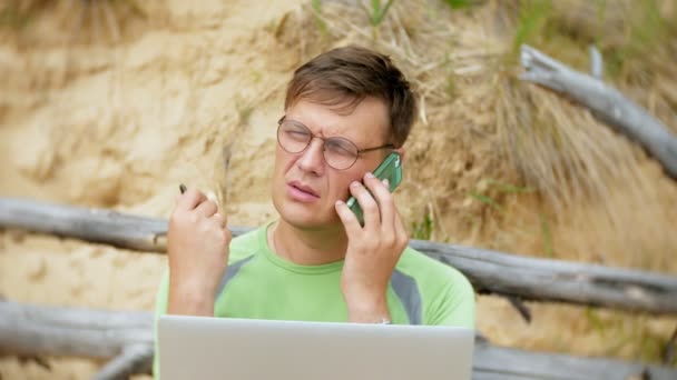 Beschäftigter älterer Mann, der an einem Laptop arbeitet, auf dem Geschäftspapiere liegen und zum Telefon greift, um an einem sonnigen Tag am Strand am Meer zu telefonieren und Wein aus einem Glas zu trinken — Stockvideo