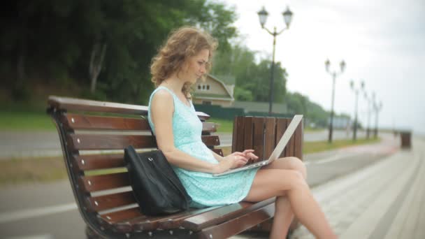 En kvinna sitter på en bänk på stranden med en bärbar dator — Stockvideo