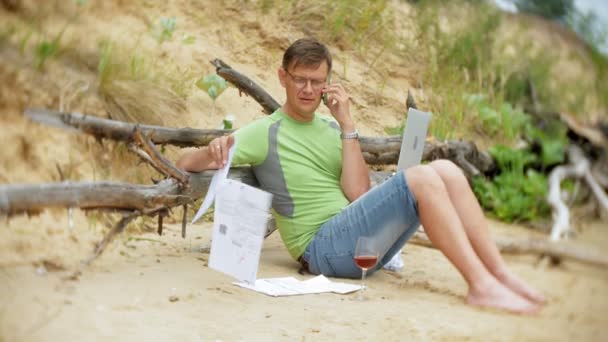 Зайнятий зрілий чоловік працює на портативному комп'ютері з бізнес-документами, підбираючи телефон для роботи дзвінка, що сидить на пляжі біля моря в сонячний день і п'є вино з келиха — стокове відео
