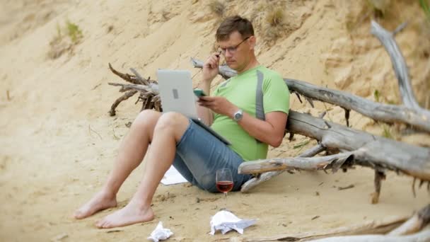 Зайнятий зрілий чоловік працює на портативному комп'ютері з бізнес-документами, підбираючи телефон для роботи дзвінка, що сидить на пляжі біля моря в сонячний день і п'є вино з келиха — стокове відео