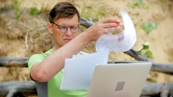 Hombre maduro ocupado trabajando en una computadora portátil con papeles de negocios recogiendo un teléfono para trabajar una llamada sentada en la playa junto al mar en un día soleado y bebiendo vino de una copa — Vídeo de stock