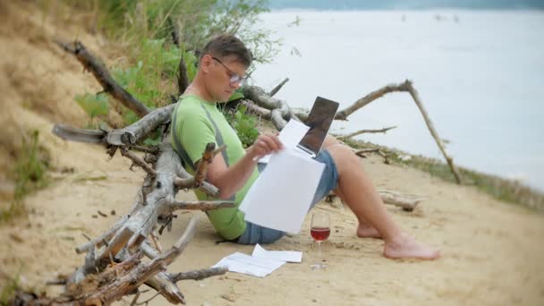 Upptagen mogen man arbetar på en bärbar dator med affärshandlingar plocka upp en telefon att arbeta ett samtal som sitter på stranden vid havet på en solig dag och dricka vin ur ett glas — Stockvideo