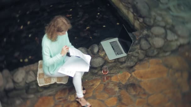 Mulher com um laptop e documentos no fundo de uma parede de pedra. Bebidas e bebidas vinho de um copo — Vídeo de Stock