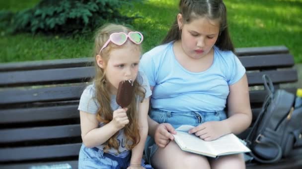 Дві маленькі дівчата сидять на дерев'яній лавці в місті, читаючи книгу і їдячи морозиво, на фоні міського парку — стокове відео