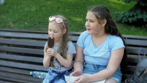 Dwie małe dziewczynki siedzą na ławeczce, drewniane, w mieście, czytając książkę i jeść lody, na tle parku miejskiego — Wideo stockowe