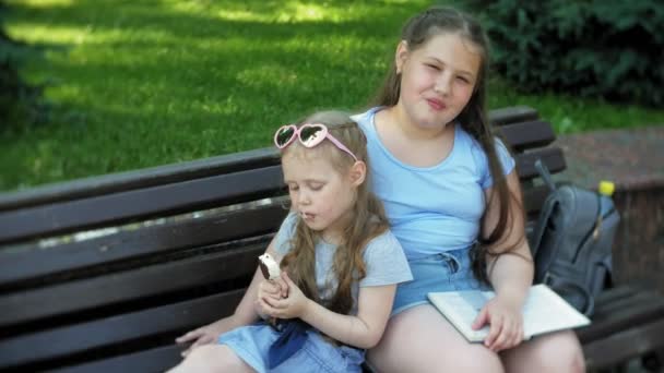 Duas meninas estão sentadas em um banco de madeira em uma cidade lendo um livro e comendo sorvete, o fundo de um parque da cidade — Vídeo de Stock