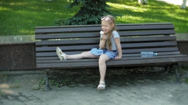 Маленькая девочка сидит на деревянной скамейке в городе, ест мороженое, на заднем плане городского парка — стоковое видео