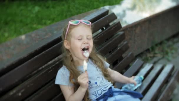 Маленька дівчинка сидить на дерев'яній лавці в місті, їсть морозиво, фон міського парку — стокове відео