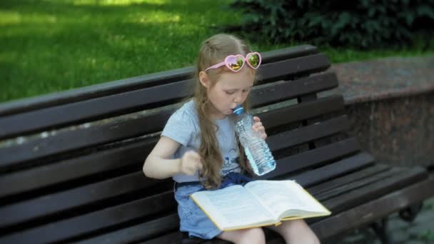 小女孩坐在一个木头长凳在城市读一本书, 城市公园的背景 — 图库视频影像