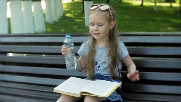 Niña sentada en un banco de madera en una ciudad leyendo un libro, fondo de un parque de la ciudad — Vídeo de stock