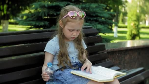 Menina sentada em um banco de madeira em uma cidade lendo um livro, fundo de um parque da cidade — Vídeo de Stock