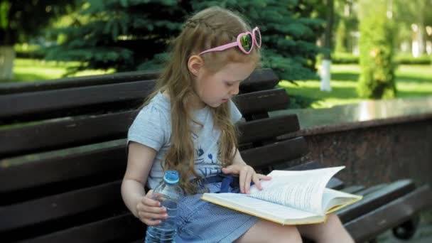 Niña sentada en un banco de madera en una ciudad leyendo un libro, fondo de un parque de la ciudad — Vídeo de stock