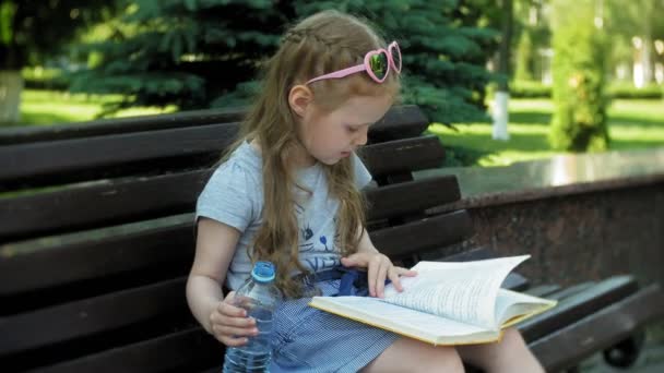 Dívka sedící na dřevěnou lavici v městě čte knihu, pozadí městského parku — Stock video