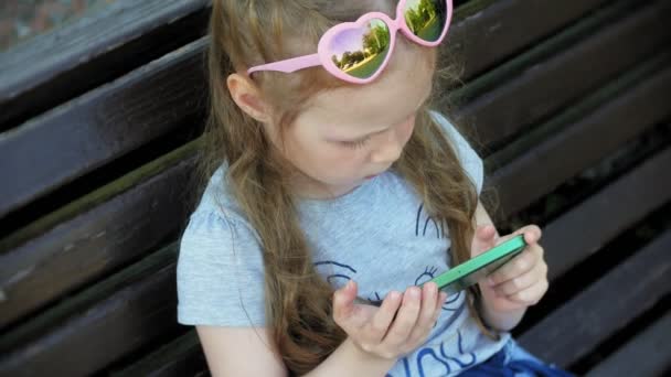 Liten flicka sitter på en träbänk i staden använder en smartphone, stadspark bakgrund — Stockvideo