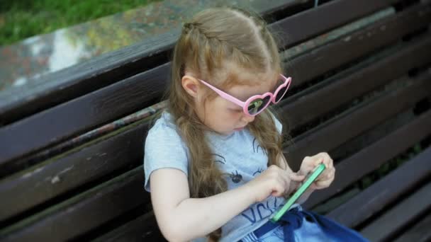 Liten flicka sitter på en träbänk i staden använder en smartphone, stadspark bakgrund — Stockvideo