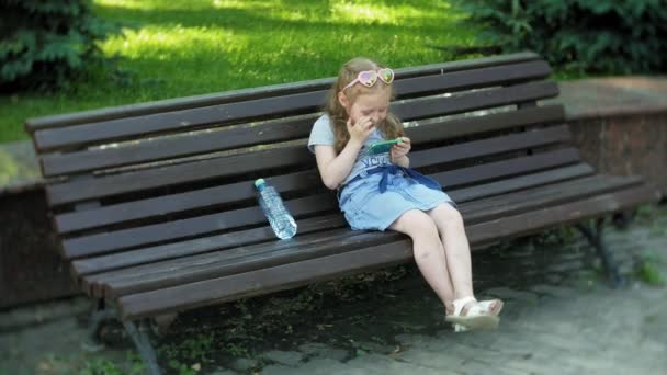 Liten flicka sitter på en träbänk i staden använder en smartphone, stadspark bakgrund — Stockvideo