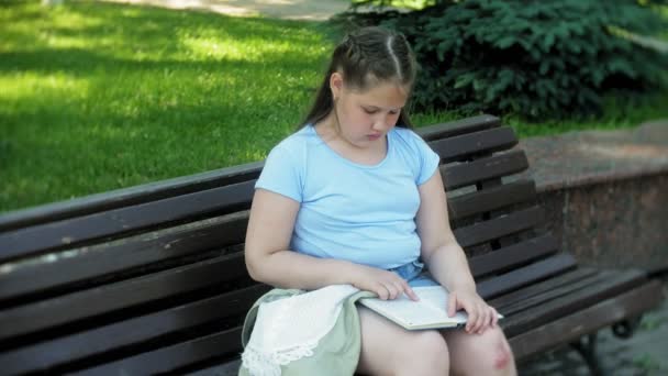 Jong meisje zittend op een houten bankje in een stad die lezen van een boek, de achtergrond van een stadspark — Stockvideo