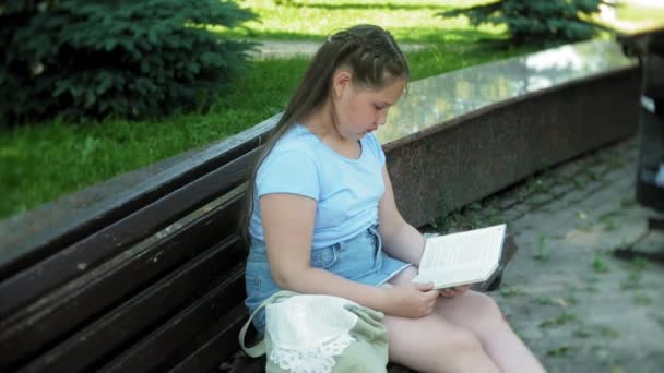 Menina sentada em um banco de madeira em uma cidade lendo um livro, fundo de um parque da cidade — Vídeo de Stock