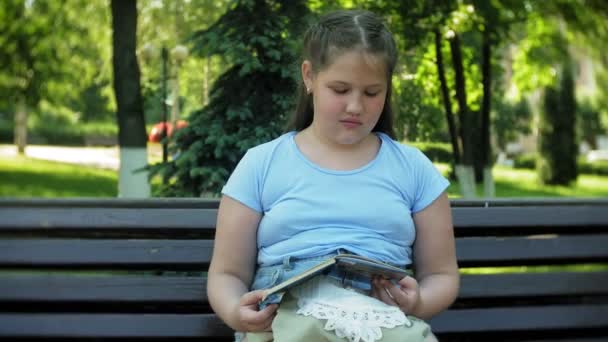 Menina sentada em um banco de madeira em uma cidade lendo um livro, fundo de um parque da cidade — Vídeo de Stock