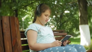Küçük bir tablet Pc ile şişman kız ve müzik gerekse video bir yaz parkta bir bankta oturan kulaklıklar