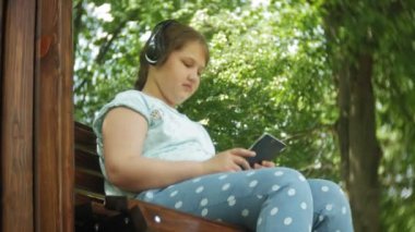 Küçük bir tablet Pc ile şişman kız ve müzik gerekse video bir yaz parkta bir bankta oturan kulaklıklar