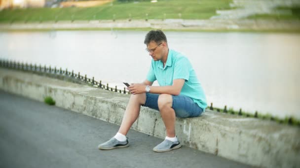 Joven hombre feliz con un teléfono inteligente en el parque de la ciudad se sienta en la orilla — Vídeos de Stock