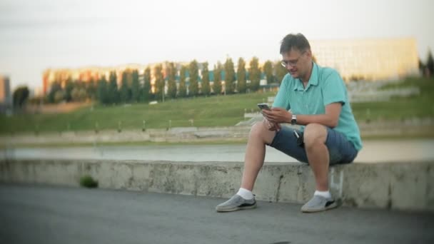 Молодой счастливый человек со смартфоном в городском парке сидит на берегу — стоковое видео