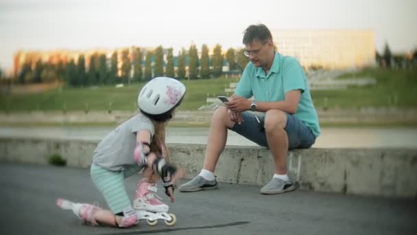 Отец и дочь катаются на роликовых коньках. Девочка учится кататься на роликах и падает. Отец учит дочь кататься на роликах — стоковое видео